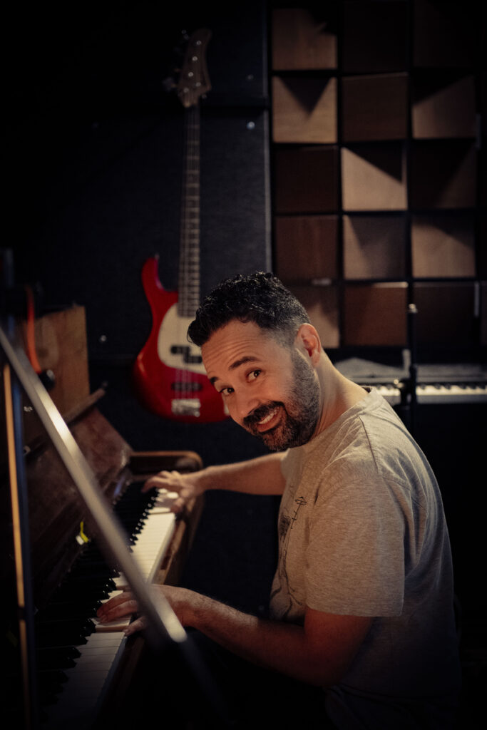 Man smiling while playing piano in a recording studio, with guitar and acoustic panels in the background.