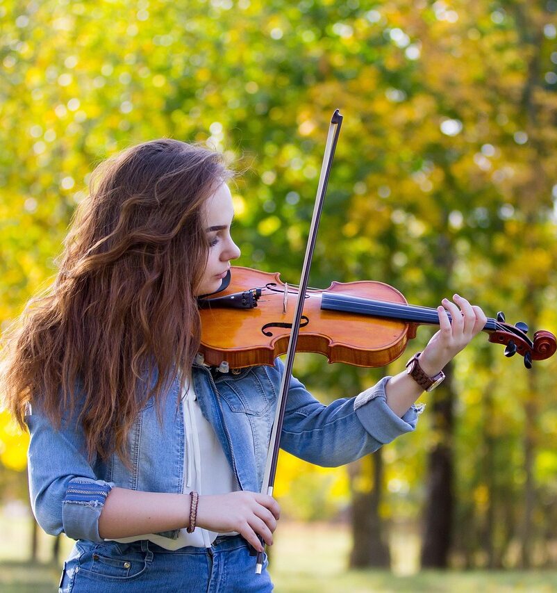 Young girl playing violin in a schoolyard with trees in the background, wearing a sweater – music lesson in nature