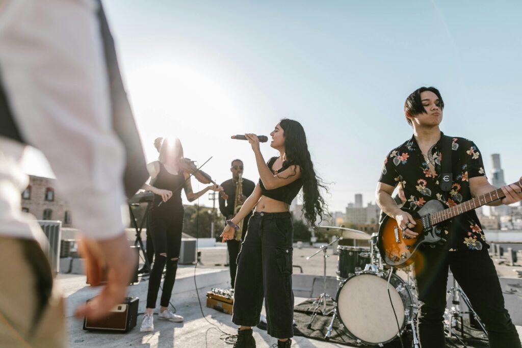 A group of students playing musical instruments outdoors, including guitar and drums, performing with energy and joy.